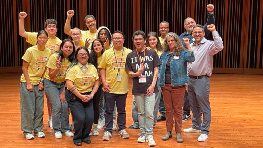 Macalester student Marco Lopez ’27 poses with a group of peers and mentors on stage, smiling and raising their fists to celebrate.