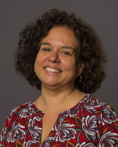 Professor Cláudia Tatinge Nascimento smiles with arms crossed in a softly lit hallway.