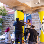 Students work together to paint colorful designs on outdoor columns as part of a community art project during Mac Fest 2025