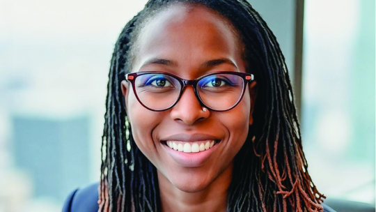 Yeukai Mudzi ’12 wearing glasses and a navy blazer smiles while seated near a window with a city skyline in the background.