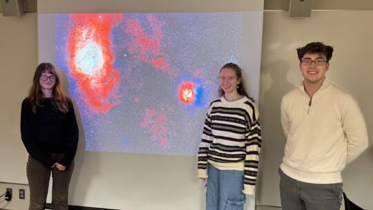 Marine McKnight '28, Emma McNellis '28, and Hayden Arko '27 pose in front of a powerpoint presentation, which displays an image of a galaxy nebula.
