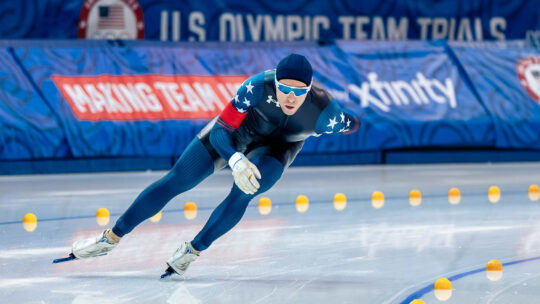 A male speedskating athlete skates around the corner of track on an ice rink.