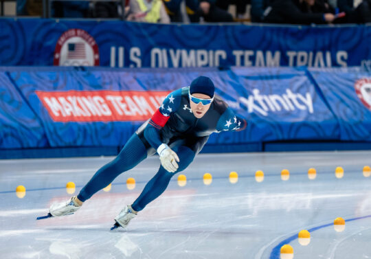 A male speedskating athlete skates around the corner of track on an ice rink.