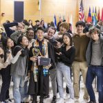 Twelve people pose for a picture with a Macalester 2022 December graduate. Many of the people are looking at the graduate and pointing to them.