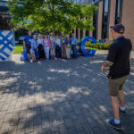 A Reunion attendee takes a picture using a cell phone of a group of nine Reunion attendees standing in front of a sculpture of the letters MAC