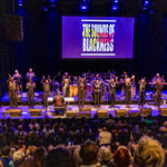 The ensemble Sounds of Blackness performs on a stage, with a screen reading SOUNDS OF BLACKNESS displayed behind them and an audience watching them