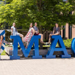 Five Reunion attendees sit on top of a sculpture reading MAC, while President Rivera stands next to the sculpture