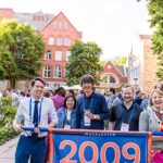 A group of five alumni stand behind a banner reading 2009