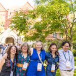 Five alumni walk toward the camera in a horizontal line, arms around each other and smiling