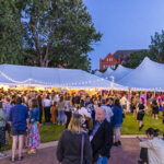 A crowd of Reunion attendees stand talking to each other under and around a tent at night