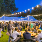 A crowd of Reunion attendees stand talking to each other under and around a tent at night