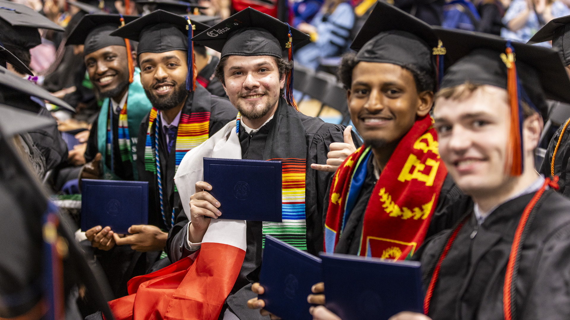 Five graduates sit in a row and smile for the camera while holding their diplomas