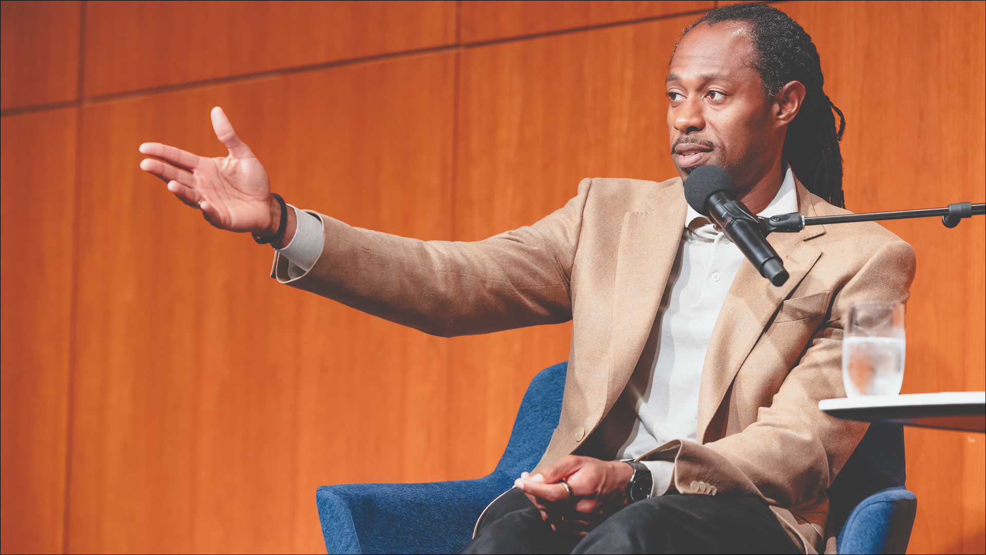 Dr. Keon West ’06 speaks into a microphone during a Reunion event, gesturing with one hand while seated on stage in a tan blazer and white shirt.