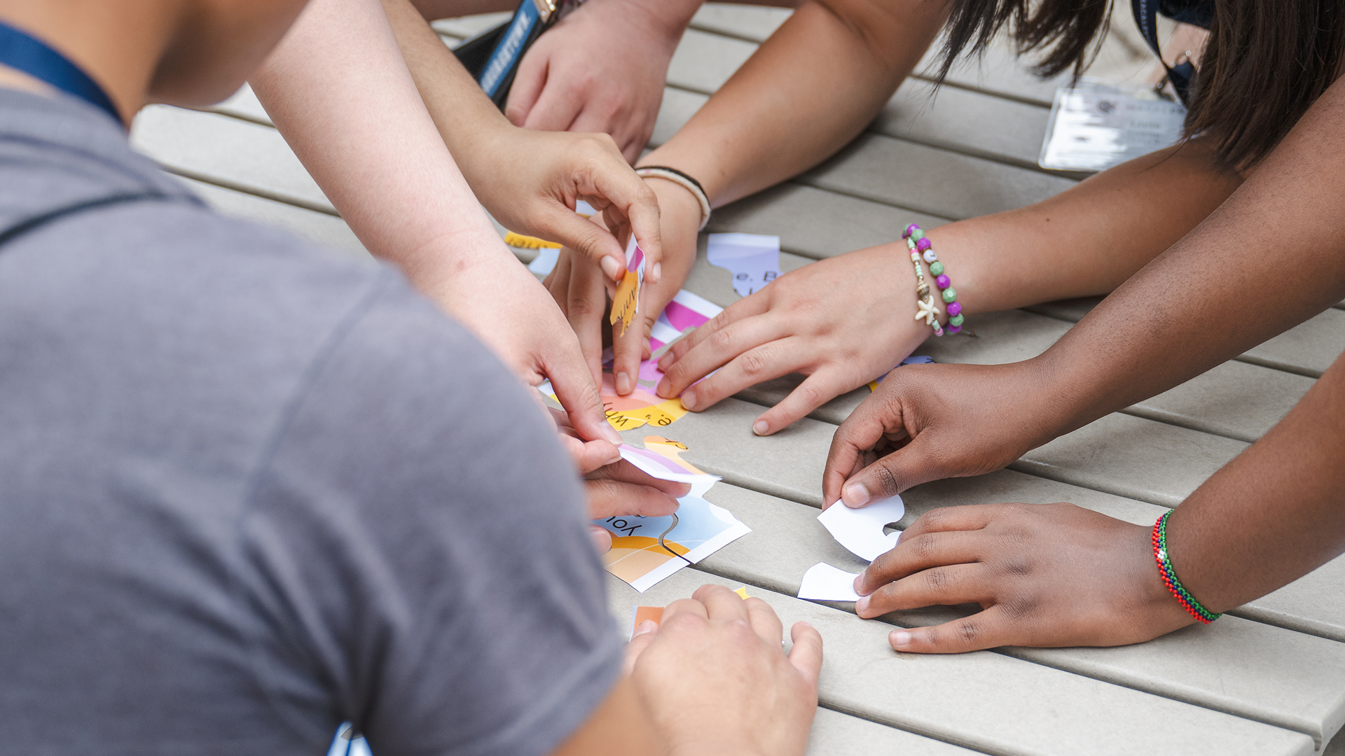 Close-up of students’ hands collaborating to piece together a colorful puzzle, symbolizing teamwork, peer teaching, and learning through shared effort.