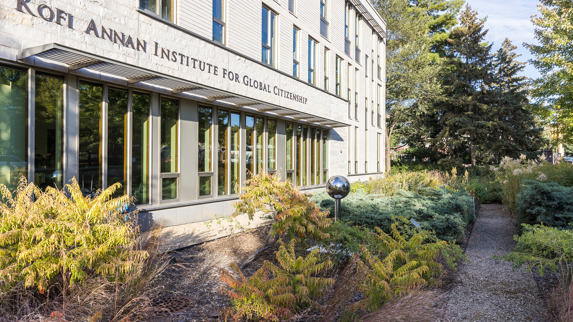 Exterior of the Kofi Annan Institute for Global Citizenship at Macalester College, with sunlight reflecting off its large windows and a landscaped garden pathway in the foreground.