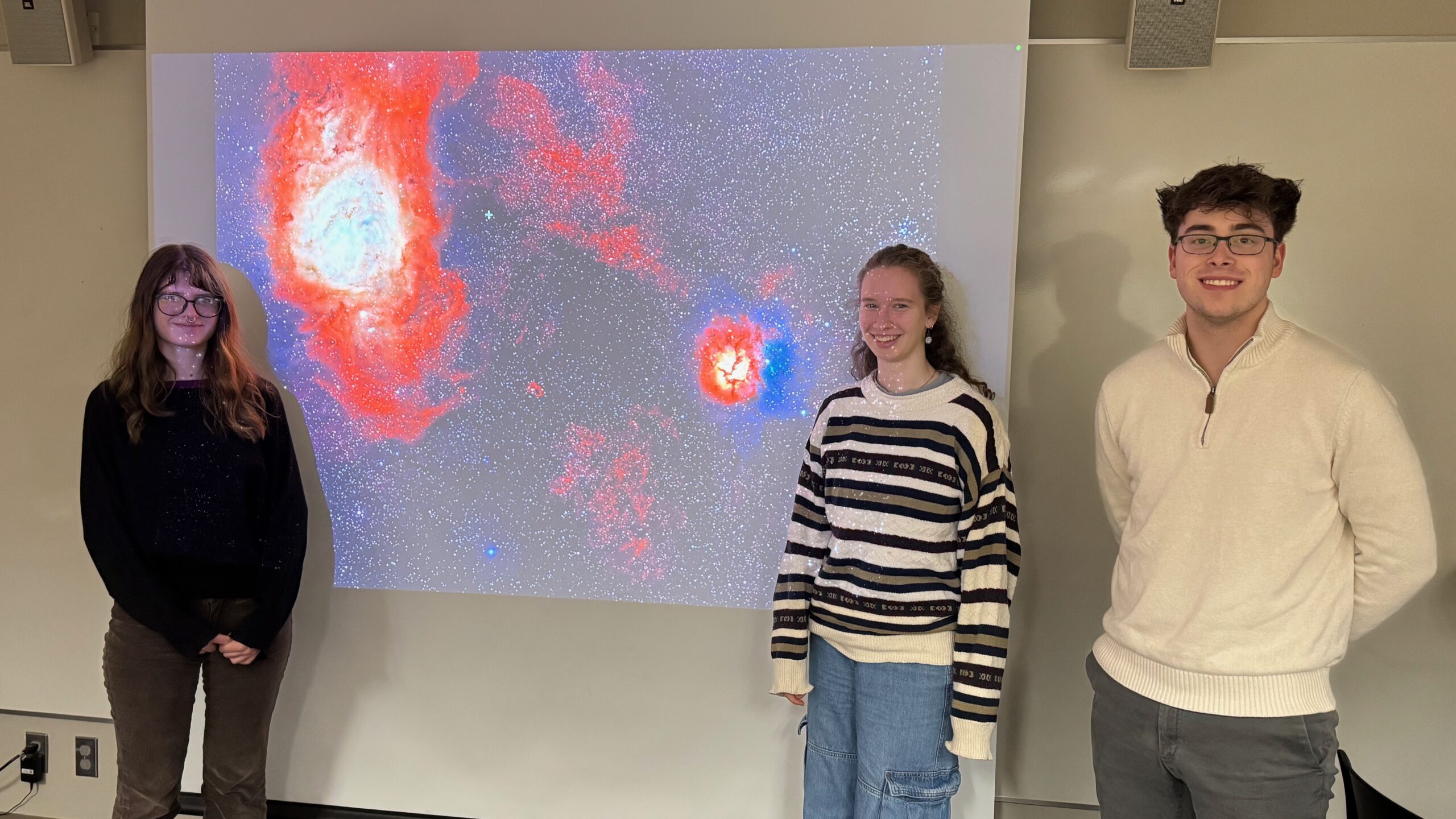 Marine McKnight '28, Emma McNellis '28, and Hayden Arko '27 pose in front of a powerpoint presentation, which displays an image of a galaxy nebula.