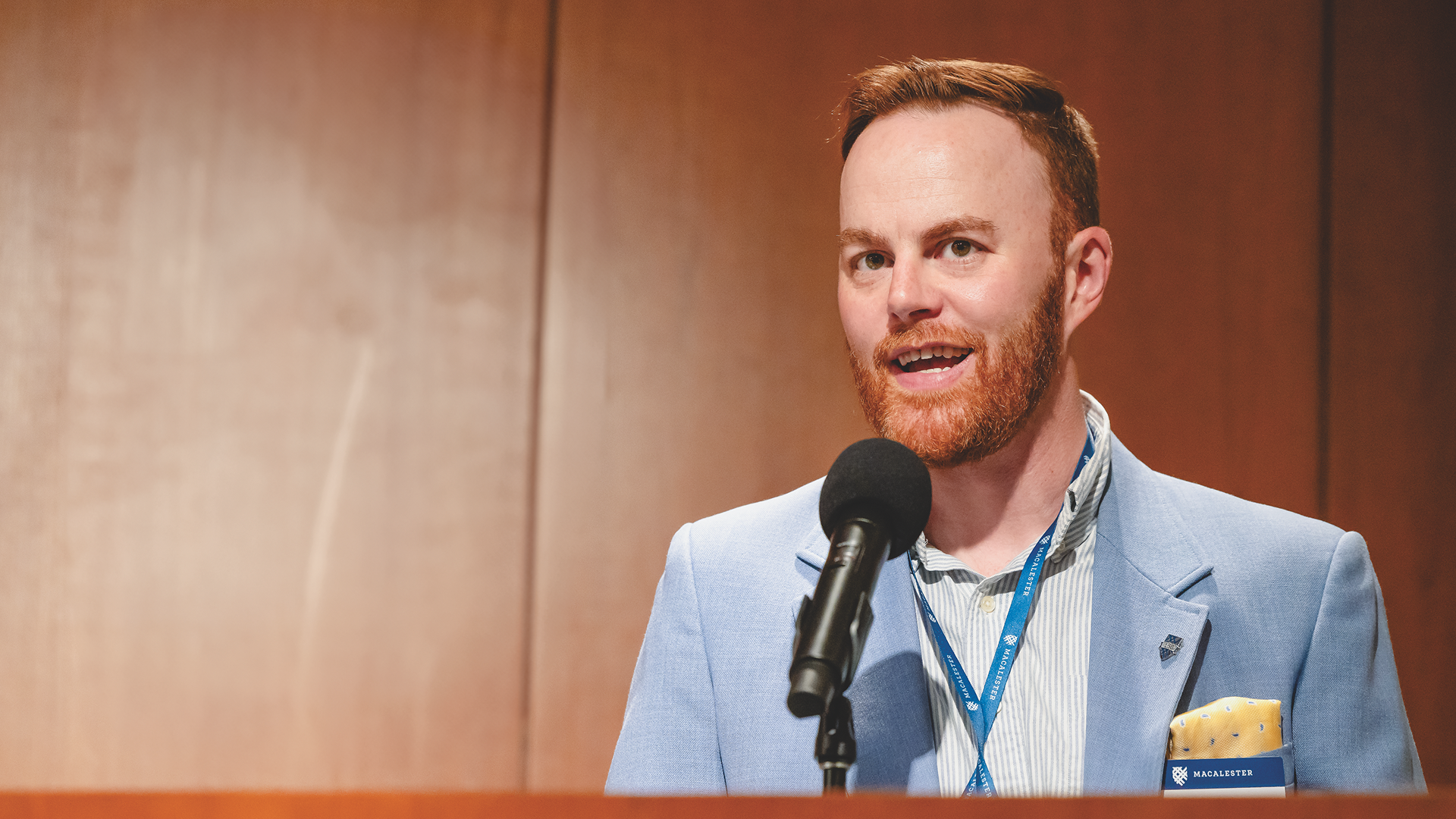 A portrait of Alumni Board President Kevin Finnegan ’11, a man with a red beard wearing a light blue blazer and a yellow pocket square, speaking into a microphone.