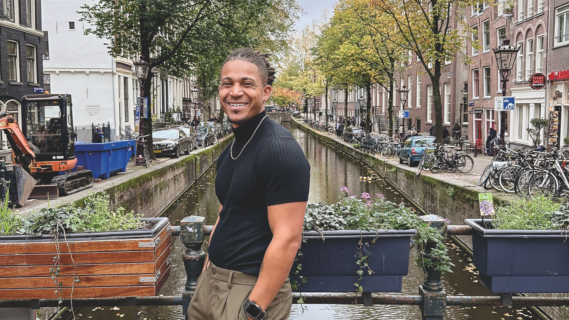 A portrait of Macalester alumnus Kiante Miles ’20, smiling while standing on a bridge over a canal in Amsterdam, wearing a black turtleneck and gold chain.