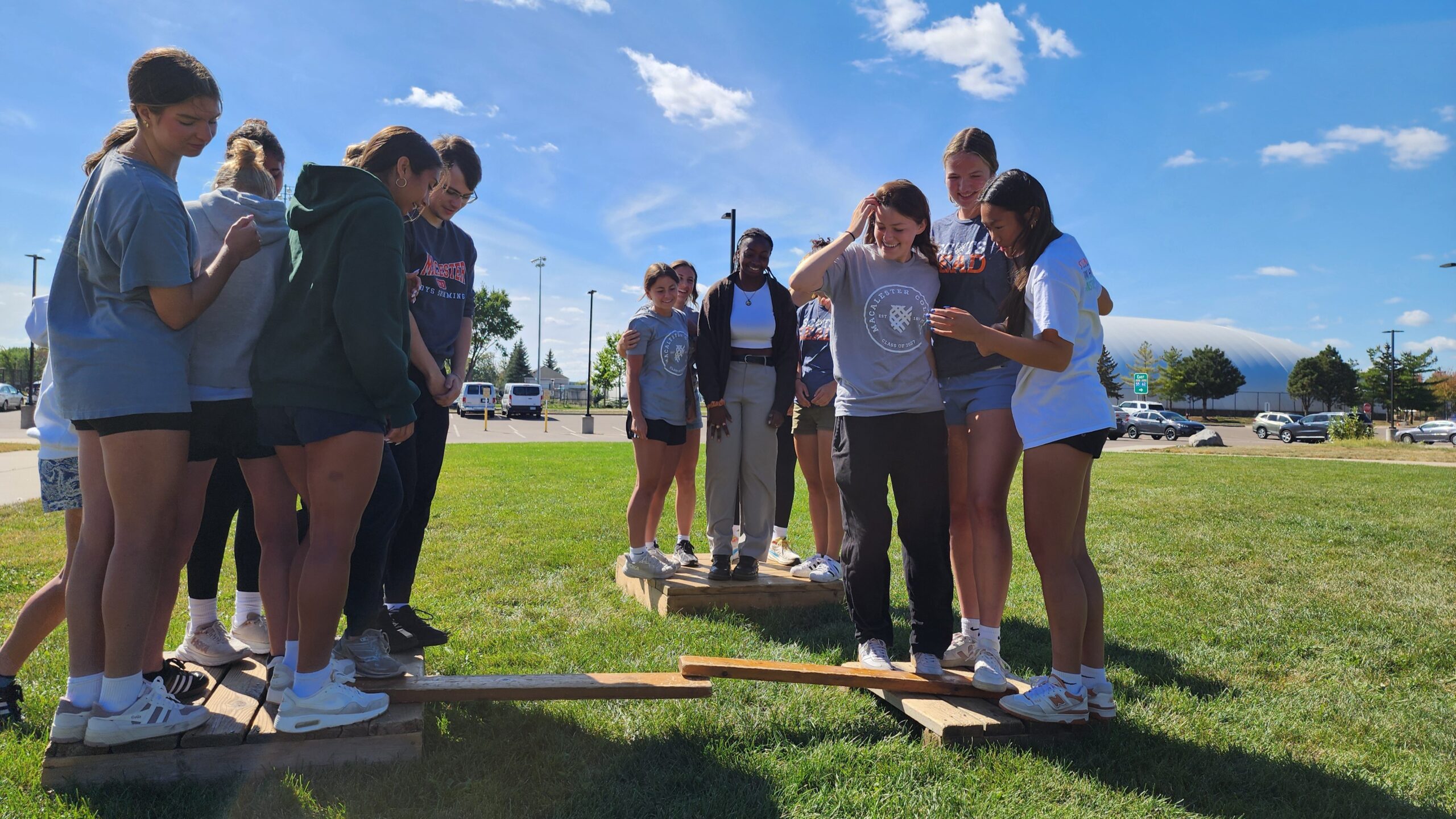Macalester students participate in a team-building activity outdoors, balancing on wooden boards during a leadership exercise.