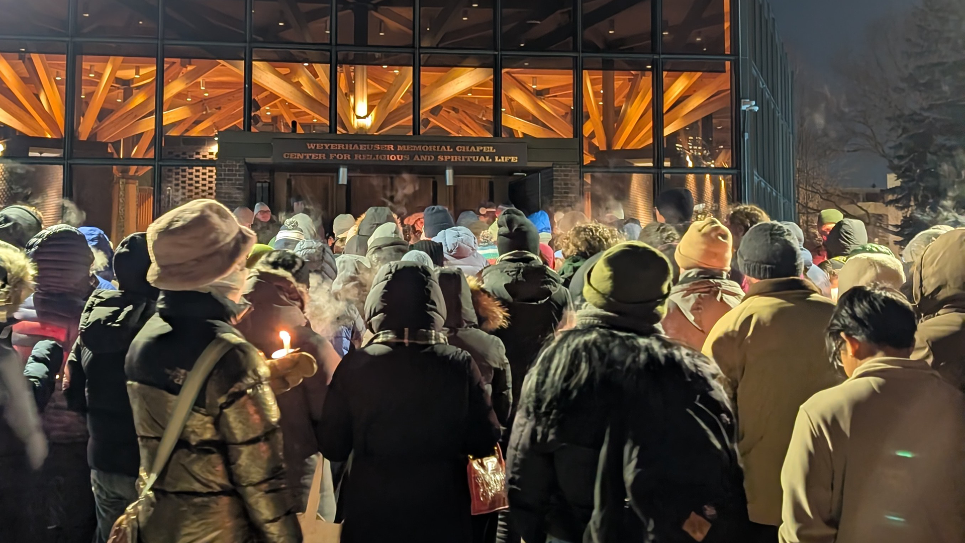 A large crowd of people in winter coats gather outside the Weyerhaeuser Memorial Chapel at night, some holding lit candles for the "Shine a Light for Minnesota" vigil.