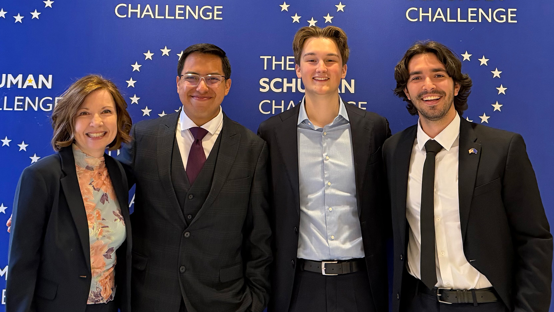 Three students and a professor stand in front of a blue backdrop that says THE SCHUMAN CHALLENGE