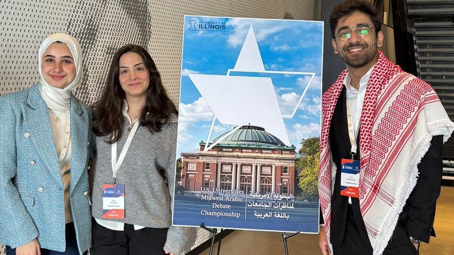 Three Macalester students pose by a poster for the U.S. Universities Midwest Arabic Debate Championship