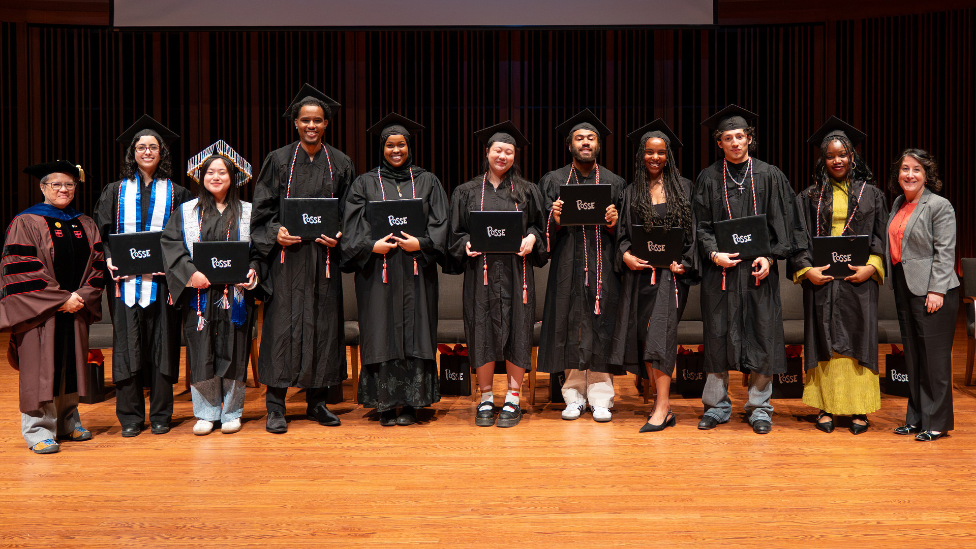 Nine Posse cohort members from the Class of 2026 wearing graduation regalia, along with President Rivera and Professor Karín Aguilar-San Juan