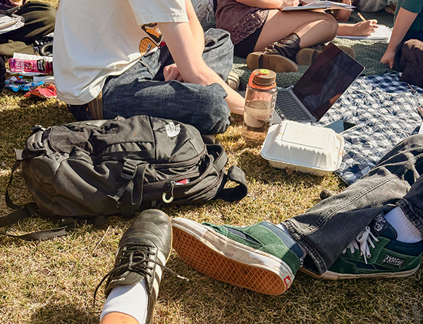 A group of students sitting on a lawn with backpacks and water bottles around them; the camera is pointing down to their legs