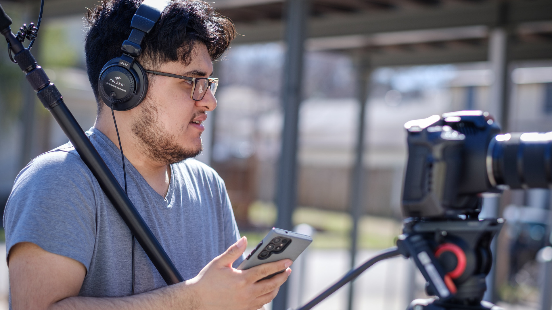 Moises Soto Puente wearing headphones and standing behind a tripod-mounted camera while holding a boom microphone