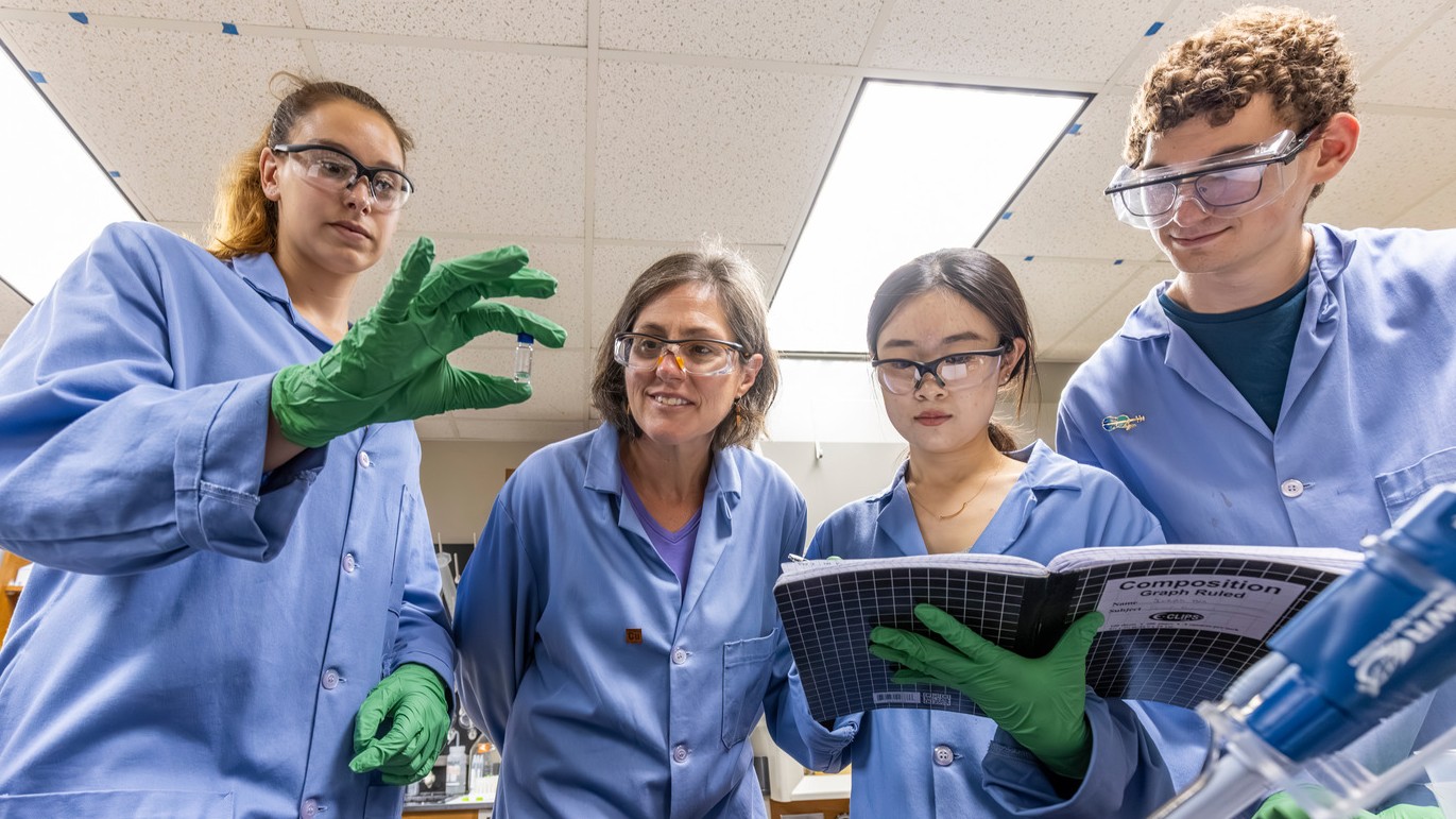 Macalester chemistry students and a professor, all wearing blue lab coats, gather around a table to look at a small vial that one student is holding. They're all wearing protective goggles, and one student is holding open a notebook.