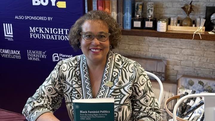 Professor Duchess Harris holds a copy of Black Feminist Politics at the Momentum Summit 2026, with event signage and shelves in the background.