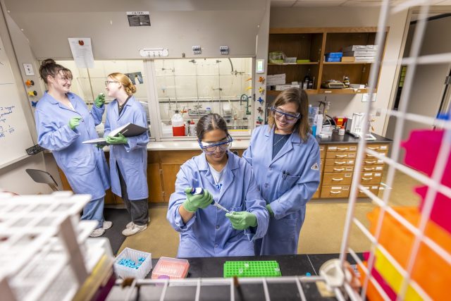 chemistry lab with students and professor in blue lab coats and safety glasses smiling