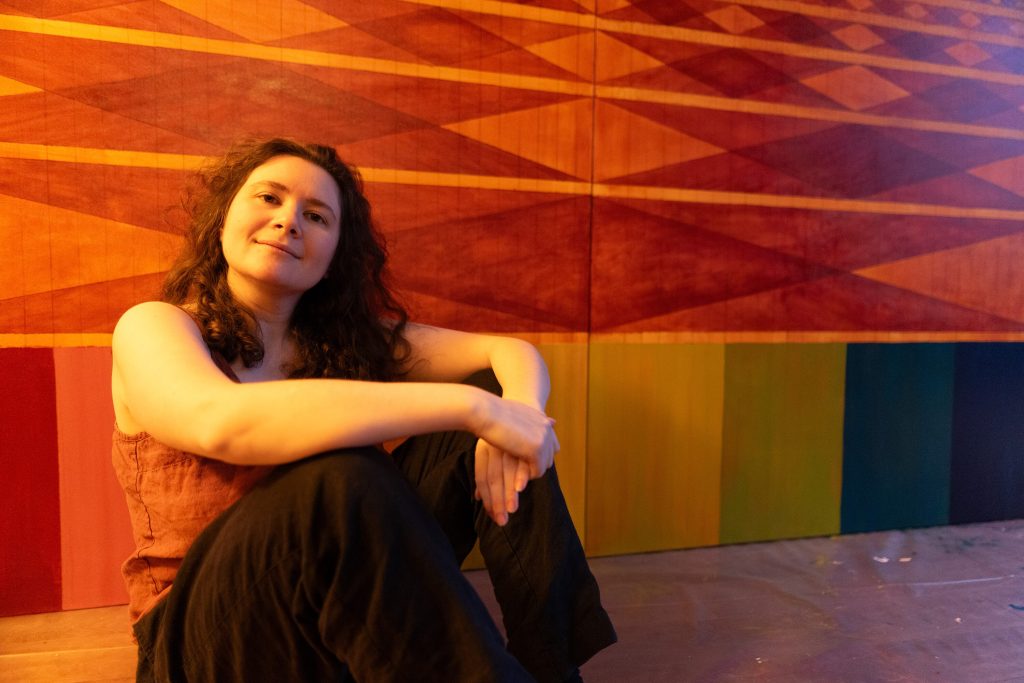 Studio portrait of artist Juliet Fiss seated in front of a large geometric painting and looking toward the camera. Angular bands of red, orange, purple, green, and gold fill the background.