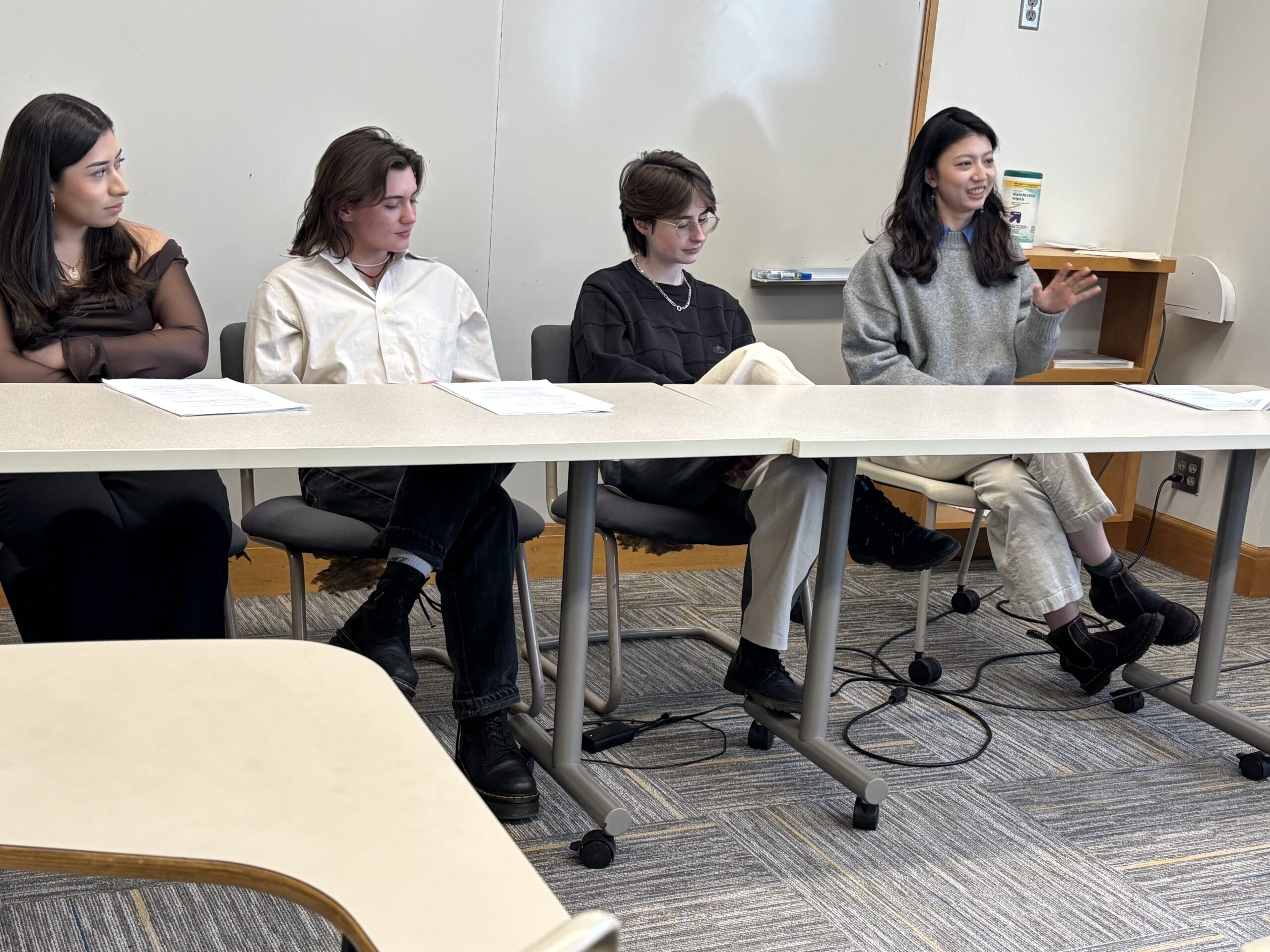 Four Macalester students sit behind a table in a Carnegie Hall classroom. The student on the right is talking, the others are listening and looking at her.