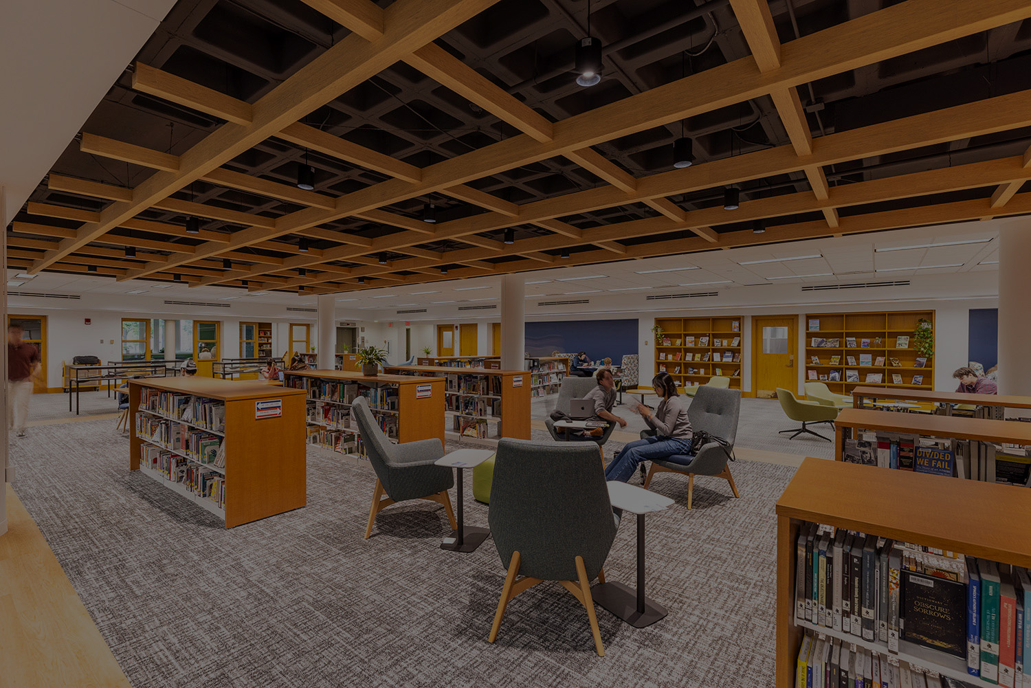 Two students sit in gray padded chairs in between six rows of waist-tall bookshelves.
