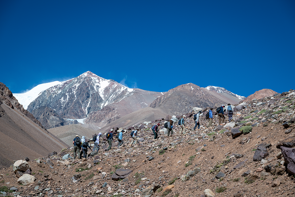 Sixteen students hike up a barren slope in the Andes Mountains. Snow-capped peak and vibrant blue sky in the background