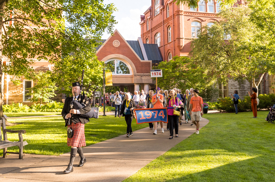 Many members of the Class of 1974 walk together under the Link, a red brick overhang with windows that connects the Library and Old Main. They are being led by a piper.