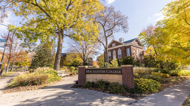 Brick Macalester sign at the entrance to campus. Trees with fall leaves and Weyerhaeuser Hall are visible in the background.