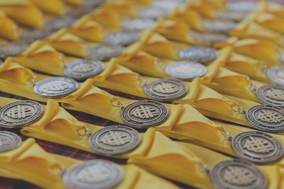 Golden Scots medals are laid on a table. The metals are brass with Macalester's shield and they are attached to yellow ribbons.