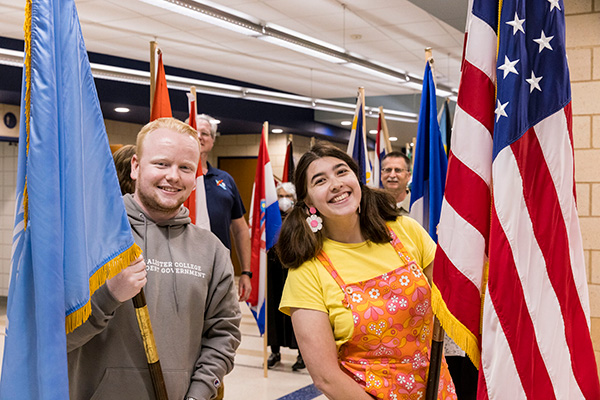 A student holding a light blue flag stands next to a student holding a United States flag