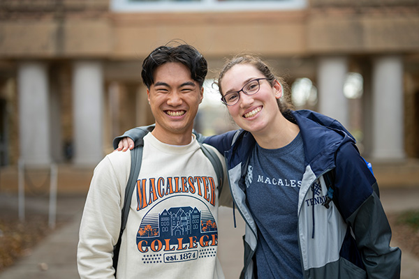 Two students wearing Macalester shirts smile and pose for the camera. One student has their hand on the other's shoulder.