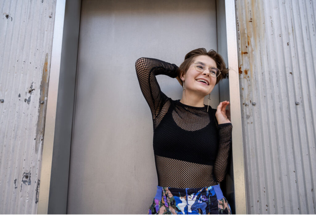 Charley Eatchel posing in front of a metal building with their arm bent at their head and smiling 