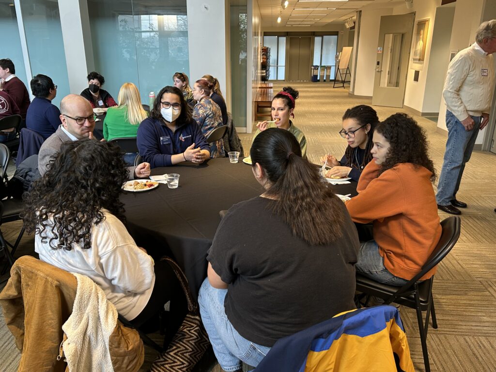 A group of alums, current students, staff and professors sit and chat at a table in Smail Gallery