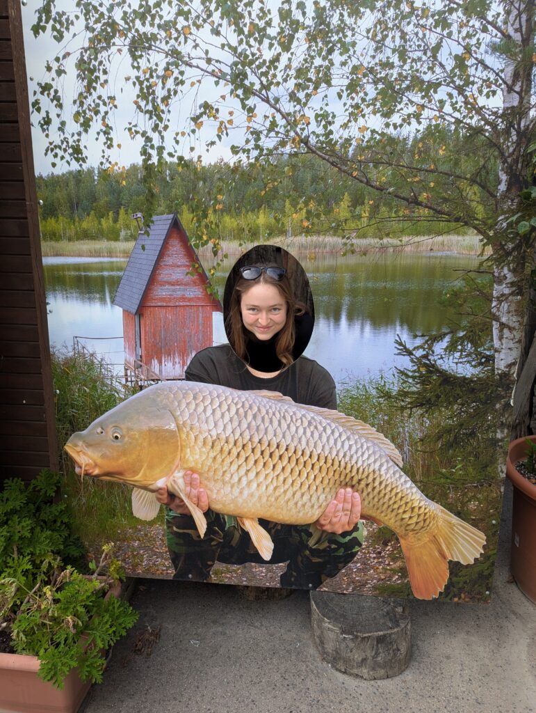 Zoe Scheuerman '24 posing with her head through a hole of a big picture of someone holding a fish by a lake.