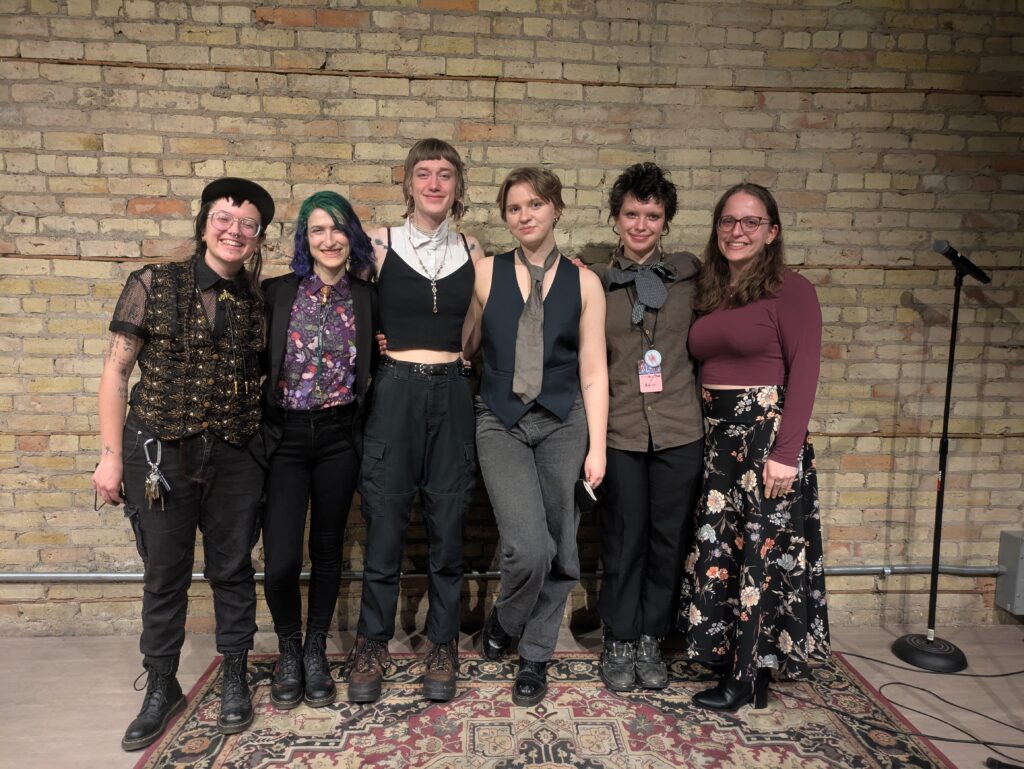 Ollie Schminkey, Natalie Kaplan, Tommy Fowler, Kendall Kieras, Adrien Wright, and Veronica Kruschel link arms to pose together for a photo on OpenBook's stage. One mic from the tournament stands to the right side.