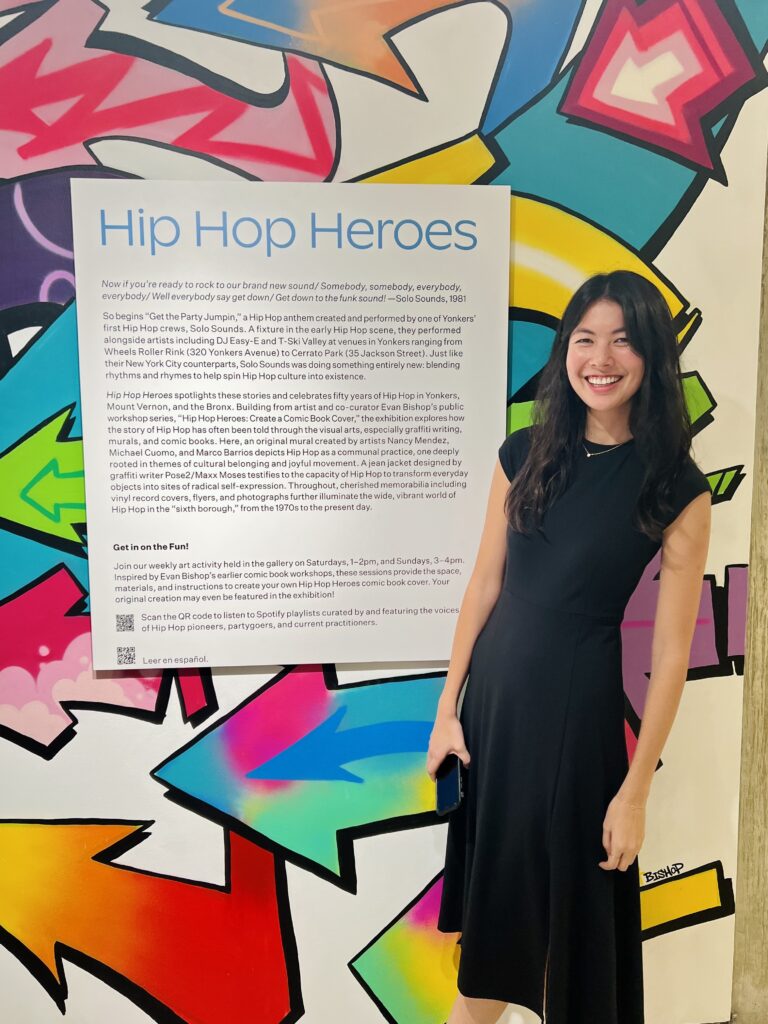 Karintha Lowe stands in front of a wall of brightly colored arrows and an exhibit summary called "Hip Hop Heroes"