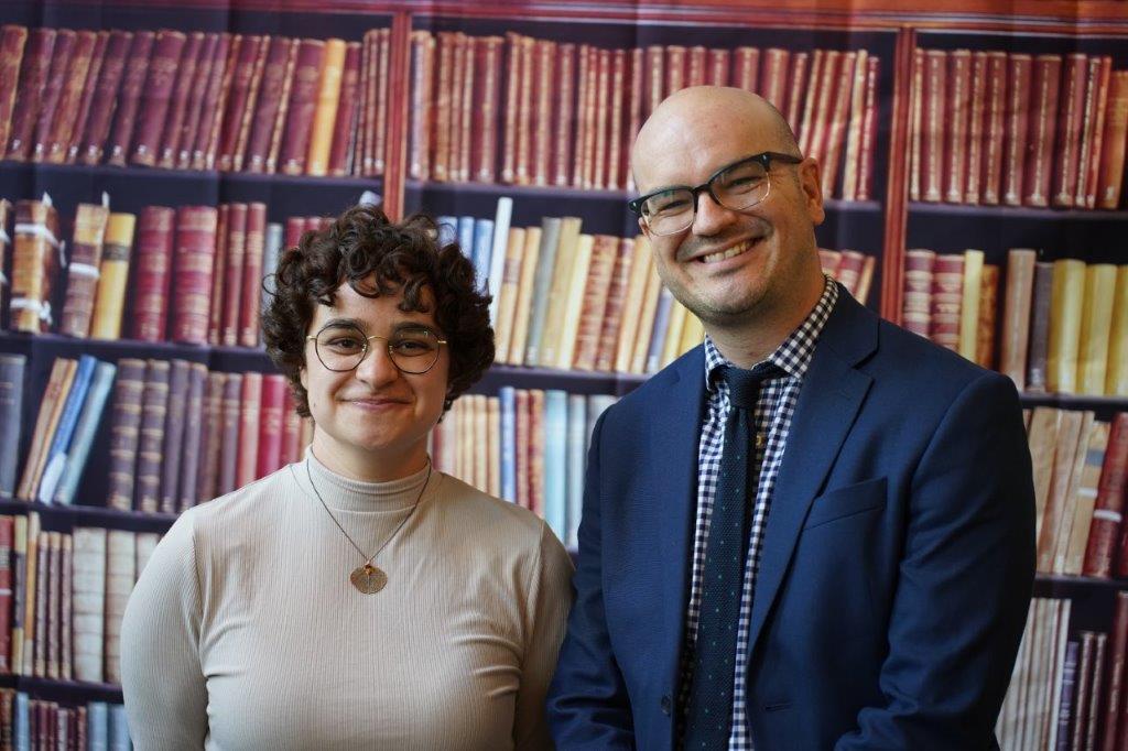 Birdie Keller stands next to Matt Burgess in front of a paper of books