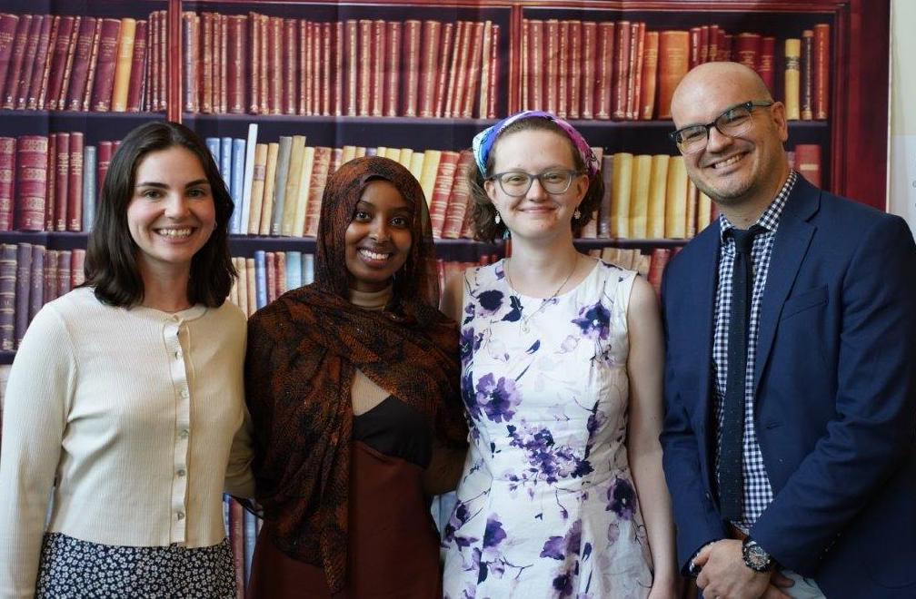 Ahlaam Abdulwali, Marley Craine, and Alma Angantyr, winners of the Wendy Parish Poetry Award, stand next to Chair Matt Burgess in front of a poster of books.