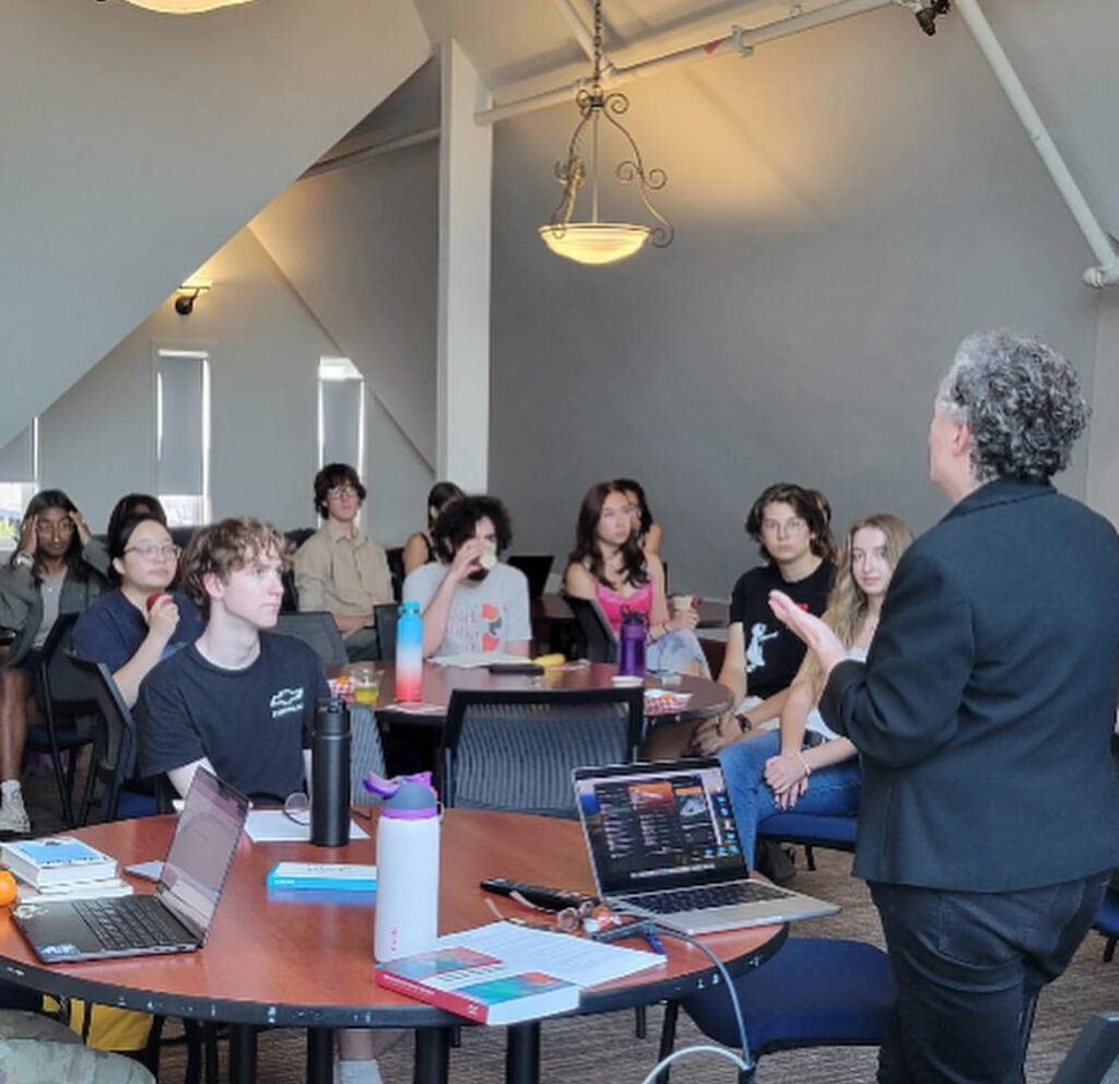 Prof. English presents before four tables of students, with her laptop open to reference songs.