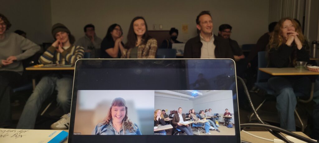 Computer screen of Zoom meeting with Megan Ritchie sits on a table in front of a crowd of smiling students (and Peter Bognanni).
